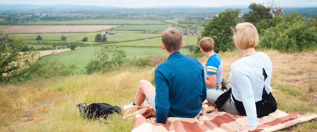 A family having a picnic on a blanket on top of a hill, looking at the view.