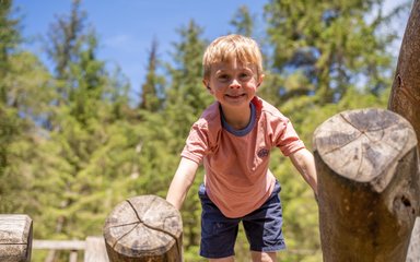 A boy on a wooden climbing frame smiling at the camera with trees in the background