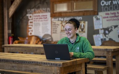 Smiling person in green fleece working a laptop on a wooden bench