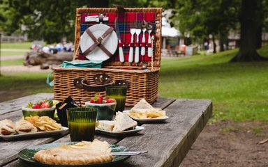 A wicker hamper with red tartan lining on a forest picnic table, shown with plates of food and drinks glasses.